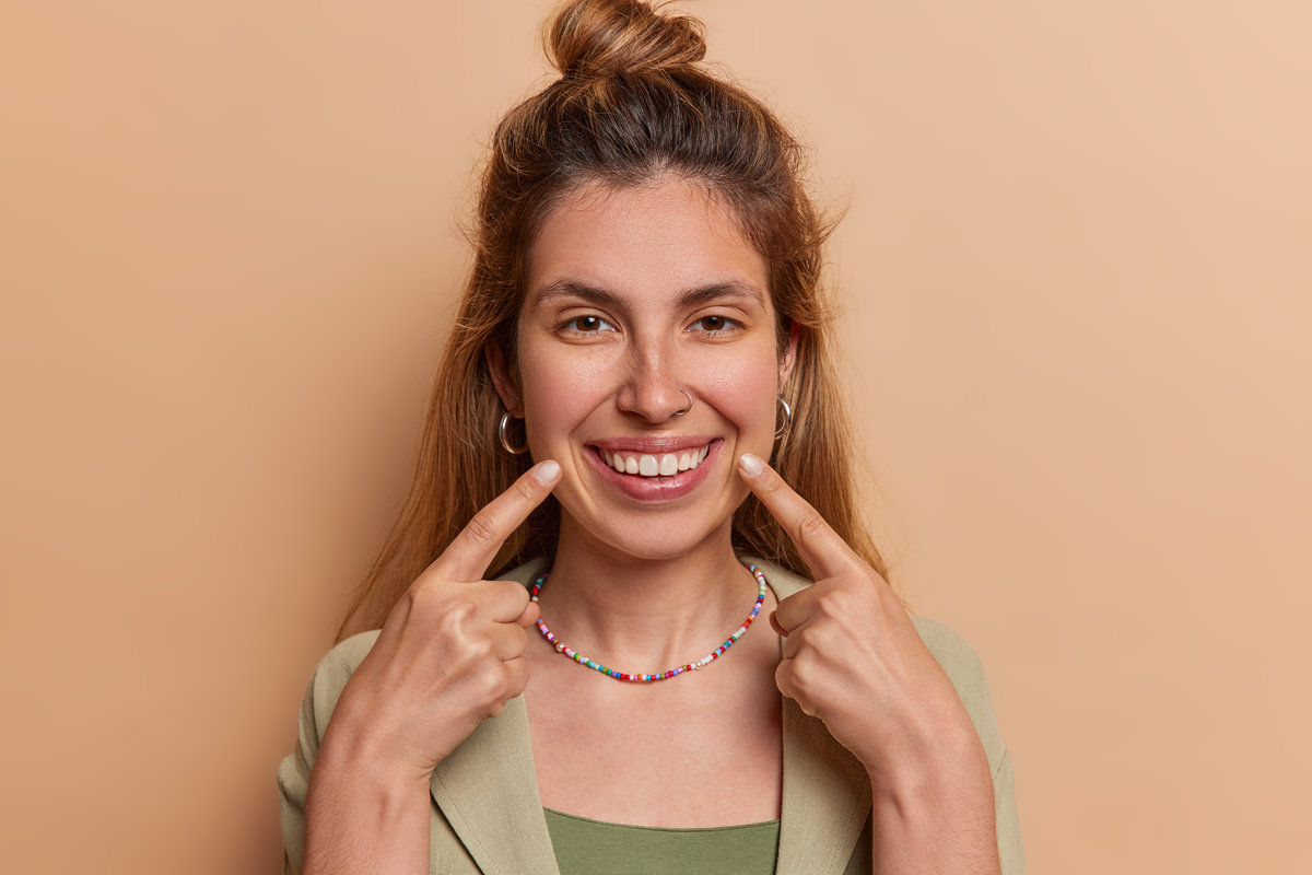Portrait of pretty European woman points index fingers at toothy smile dressed in shirt and necklace demonstrates her white teeth isolated over brown background. People and happy emotions concept