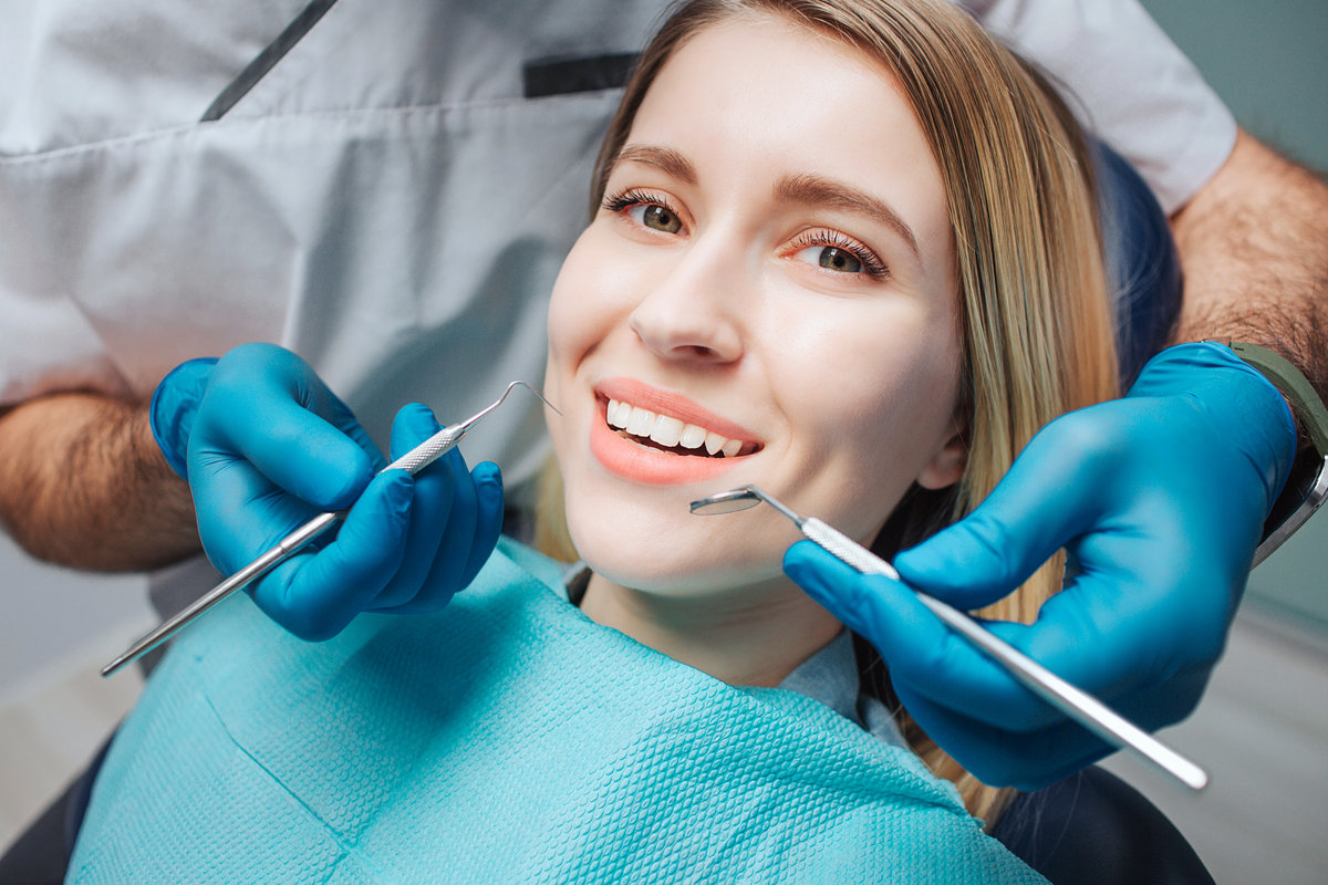 Picture of happy young woman sit in chair and look on camera. She smile and show beautiful white teeth. Doctor hold teeth tools for treatment close to mouth.