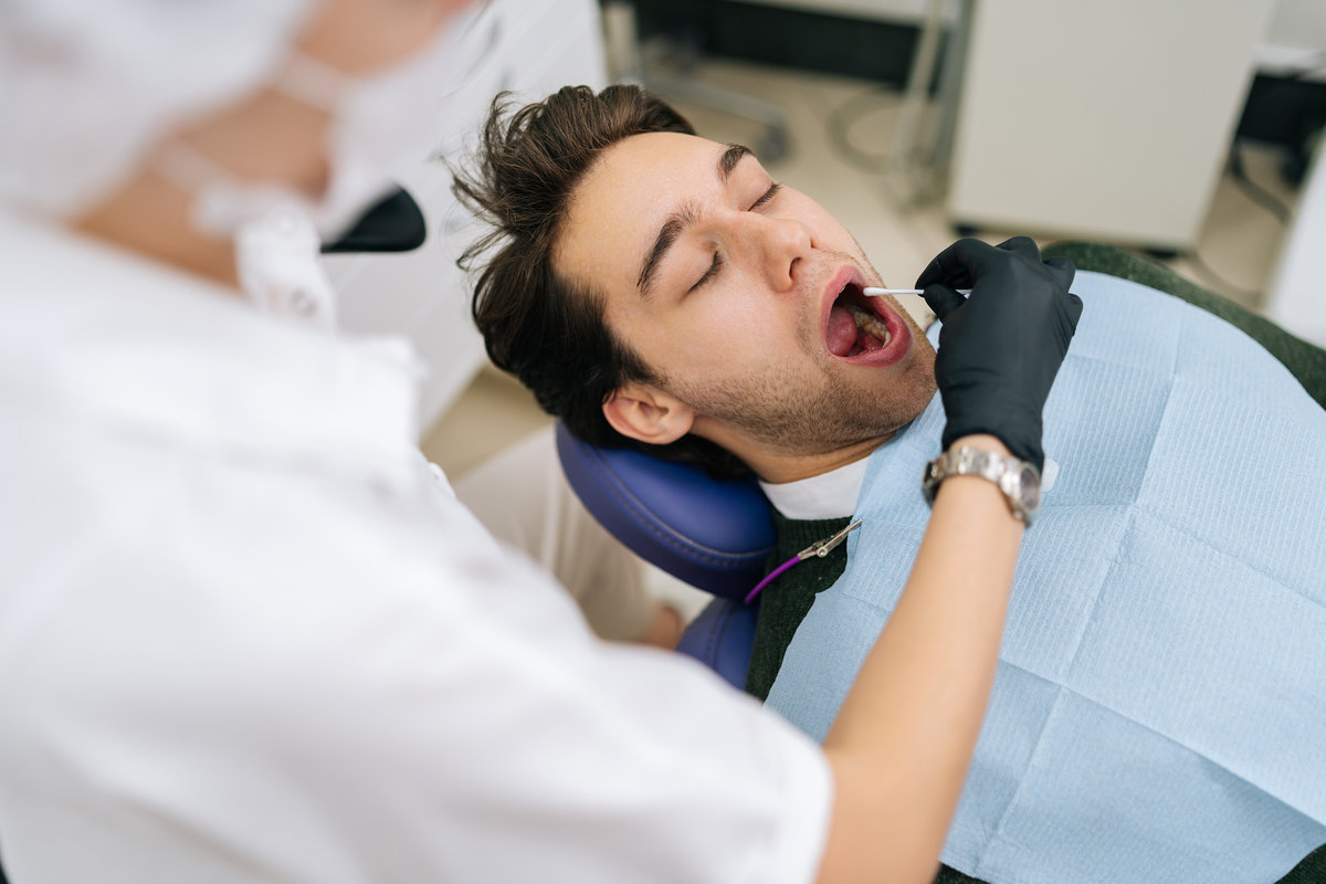 View from shoulder of unrecognizable female doctor taking saliva test from male patient mouth with cotton swab. Dentist using cotton swab to lubricate lips of man client